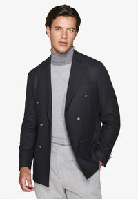 Dobbeltradet sort jakke med sølvknapper, tekstureret stof, båret over en lys grå mønstret sweater og lyse grå bukser.
