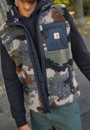 Personne portant un gilet en polaire à motif camouflage par-dessus un sweat à capuche noir, debout dehors avec un bras légèrement tendu.