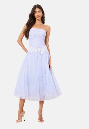 Žena nosiace fialové midi šaty bez ramienok s kvetinovou aplikáciou v páse a priehľadnú sukňu s bodkami, v kombinácii s vysokými opätkami s tenkými remienkami.