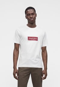 Άνδρας που φοράει λευκό T-shirt με κόκκινο ορθογώνιο και κείμενο "Jack & Jones Copenhagen", συνδυασμένο με καφέ παντελόνι, στέκεται μπροστά σε απλό φόντο.