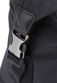 Cinghia di uno zaino nera con un'etichetta in pelle testurizzata e una fibbia in metallo lucido, su uno sfondo di tessuto liscio e dettagli in nylon.