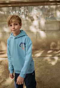 Hellblauer Kapuzenpullover mit einer Fronttasche und einem lächelnden Grafikmotiv in Orange und Lila auf der Brust, aus weichem Stoff gefertigt.