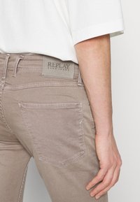 Gros plan d'une personne portant un jean Replay Blue Jeans beige avec une poche arrière et une étiquette, associé à une chemise blanche à manches courtes.
