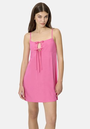 Mujer con cabello castaño liso que lleva un vestido mini rosa sin mangas, con dos lazos al frente y tirantes finos en los hombros.