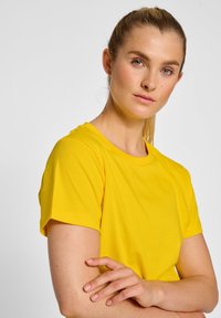Geel T-shirt met een ronde hals en korte mouwen. Zachte stof, losse pasvorm en gladde textuur. Model poseert met gekruiste armen.