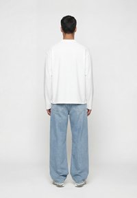 Άνδρας με την πλάτη, φοράει απλό λευκό μακρυμάνικο μπλουζάκι, ανοιχτόχρωμο τζιν παντελόνι με φαρδιά μπατζάκια και λευκά αθλητικά παπούτσια πάνω σε λευκό φόντο.