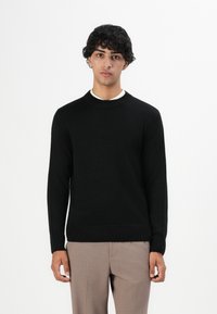 Jonge man met krullend haar, gekleed in een zwarte trui over een wit shirt en beige broek, staand voor een effen witte achtergrond.