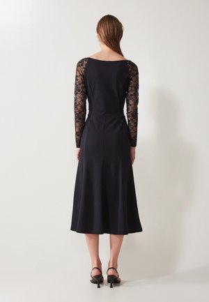 Vrouw die met haar rug naar de camera staat, gekleed in een zwarte midi-jurk met kanten lange mouwen en zwarte elegante sandaaltjes met hakken, staand tegen een effen achtergrond.