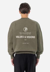 Sudadera verde oliva con un corte holgado. Presenta texto blanco en la parte trasera: logotipos y frases motivacionales. Dobladillo y puños acanalados.