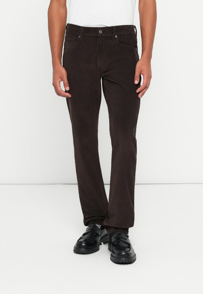 Pantalones de pana marrones con un corte recto, que cuentan con un cierre de botón y bolsillos frontales, combinados con zapatos negros de tipo slip-on.