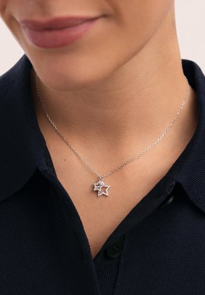 Collar de cadena plateada con un colgante en forma de estrella, adornado con pequeñas gemas transparentes, llevado sobre una camisa oscura con cuello.