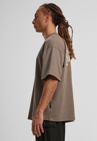 Př oversized šedé bavlněné tričko s krátkým rukávem, které má decentní logo na zádech a malou boční cedulku. Hladká textura.