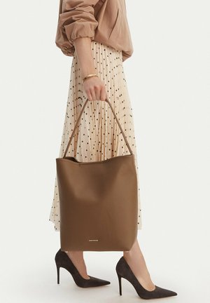 Donna con una grande borsa tote in pelle marrone, indossa una gonna beige a pois, una camicetta color cuoio, un braccialetto d'oro e scarpe con tacco nero.