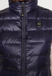 Granatowa pikowana kamizelka z wysokim kołnierzem, poziomym przeszyciem, przednim zamkiem błyskawicznym i naszywką Blauer. Gładki, błyszczący materiał o pikowanej fakturze.