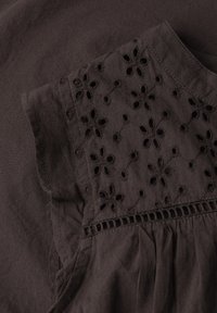 Tissu brun foncé avec broderie florale ajourée sur la partie supérieure d'un vêtement à manches courtes près de l'épaule et de l'encolure.