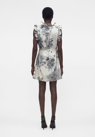 Γυναίκα με αμάνικο, βολάν, φλοράλ φόρεμα με ζώνη, στέκεται με την πλάτη γυρισμένη, φοράει μαύρα ψηλοτάκουνα μποτάκια μέχρι τον αστράγαλο.