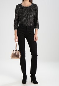 Schwarzes Glitzertop mit 3/4-Ärmeln und rundem Ausschnitt, kombiniert mit schwarzen Hosen. Mit einer metallischen roségoldenen Handtasche accessoriert.