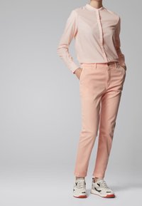 Chemise longue à manches longues rose clair avec un col mandarin, associée à un pantalon rose clair assorti, et des baskets blanches avec des accents noirs et des semelles rouges.