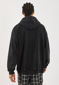 Black oversized hoodie with a hood, made of soft fabric, featuring a ribbed cuff and hem. The back shows a smooth texture, worn with checkered pants.