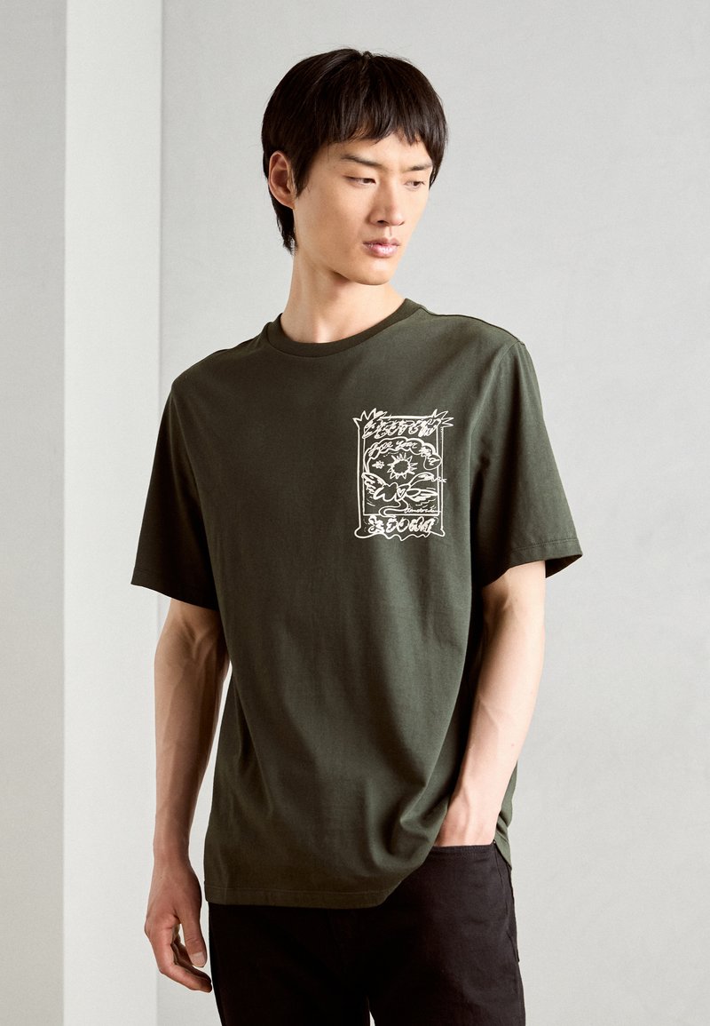 Olivengrønt bomulds T-shirt med en afslappet pasform, der har et hvidt grafisk design på venstre brystområde. Korte ærmer og rund hals.