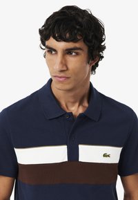 Granatowa koszulka polo z mieszanki bawełny z białymi i brązowymi poziomymi paskami. Posiada kołnierzyk, dwa guziki oraz zieloną wyszywaną wizytówkę na klatce piersiowej.