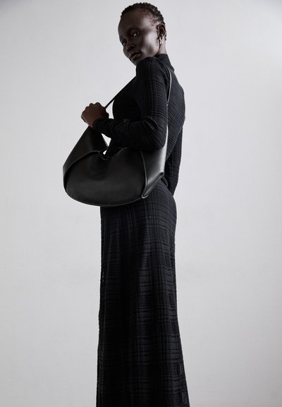 Bolso de hombro de piel negra con un diseño redondeado y costuras prominentes. Lleva un vestido negro de manga larga con textura.