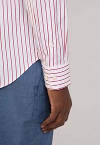 Chemise blanche avec des rayures verticales rouges, poignet boutonné, ourlet arrondi, avec un pantalon texturé bleu partiellement visible en bas.