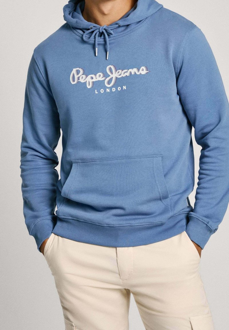 Muž oblečený v světle modré mikině Pepe Jeans London a béžových kalhotách s rukama v kapsách před jednobarevným pozadím.