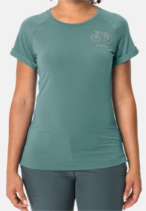 Vrouw draagt een groen T-shirt met korte mouwen en opgerolde manchetten en een kleine fietsafbeelding met het woord "now" op de bovenste linkerborst.