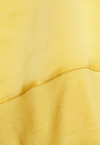Primo piano di un tessuto giallo con una texture liscia e una finitura leggermente lucida, che mette in evidenza cuciture a contrasto lungo l'orlo.