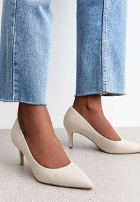 Décolleté beige in tessuto testurizzato, con punta appuntita e tacco sottile, abbinati a jeans cropped blu chiaro.