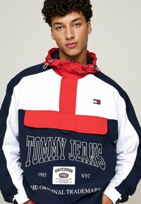 Cortavientos en navy, blanco y rojo con un bolsillo frontal con cremallera, capucha ajustable y un gran logo de "TOMMY JEANS". Fabricado con material ligero.