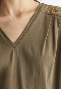 Olivově zelený top s V-výstřihem z jemné bavlny. Obsahuje šikmé ramenní švy se zlatými hroty pro přidaný detail.