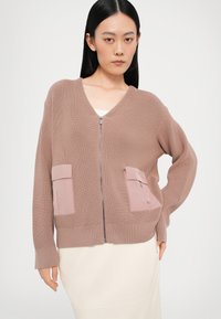 Lys brun strikket cardigan med V-hals, glidelås i midten og to frontlommer i en litt mørkere rosa nyanse. Strukturert stoff; avslappet passform.