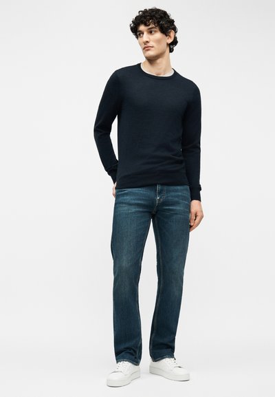 Νέος άνδρας με σγουρά μαλλιά, φοράει μαύρο πουλόβερ με μακριά μανίκια, μπλε τζιν και λευκά αθλητικά παπούτσια, στέκεται μπροστά σε απλό φόντο.