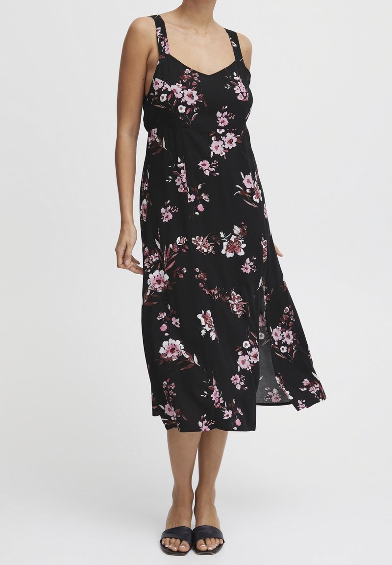 Femme portant une robe midi noire sans manches avec des motifs floraux roses et une fente latérale, associée à des ballerines noires à bouts ouverts.
