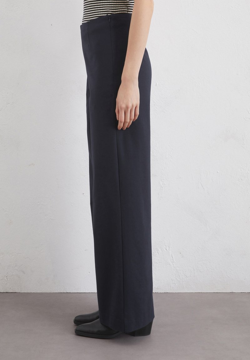 Marineblaue weit geschnittene Hosen aus Stoff mit glatter Textur, mit einem seitlichen Reißverschluss und tailliertem Schnitt, kombiniert mit spitzen schwarzen Schuhen.