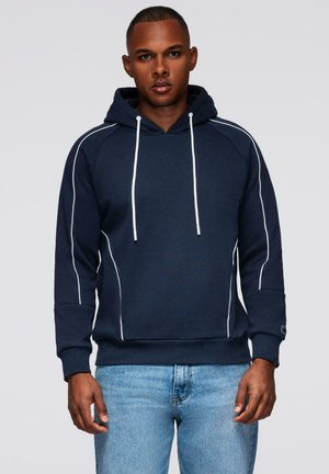 Mężczyzna w granatowej bluzie z kapturem z białymi sznurkami i detalami w jasnym odcieniu, w połączeniu z jasnoniebieskimi dżinsami, stojący na szarym tle.