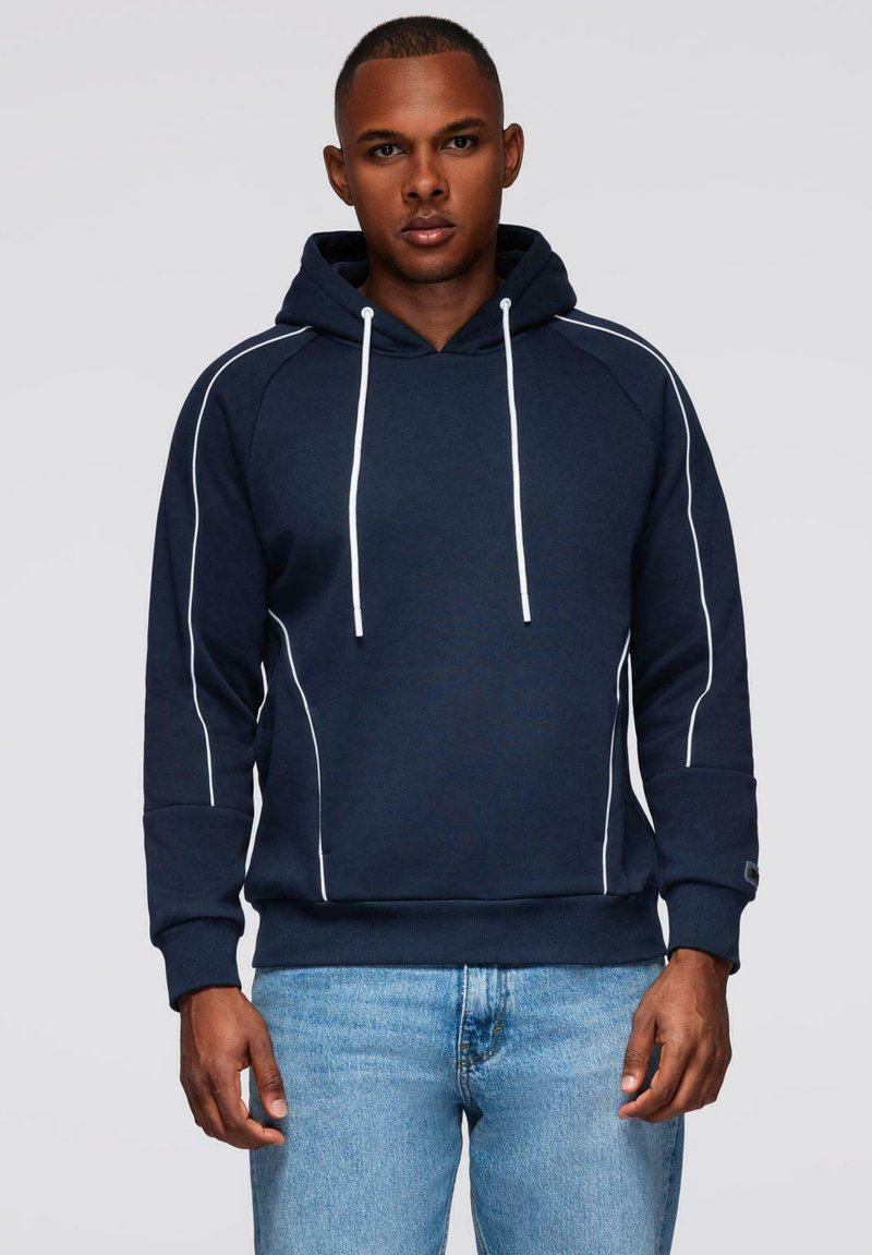 Mężczyzna w granatowej bluzie z kapturem z białymi sznurkami i detalami w jasnym odcieniu, w połączeniu z jasnoniebieskimi dżinsami, stojący na szarym tle.