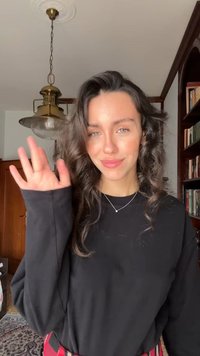 Czarna bluzka z długim rękawem o luźnym kroju, zestawiona z czerwono-czarnymi spodniami w kratę. W tle półki z książkami i sufitowa lampa.