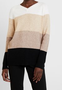 Maglione a V con strisce orizzontali in bianco, beige, marrone chiaro e nero. Realizzato in un tessuto morbido con polsini e orlo a coste.