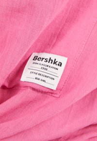 Biała metka na różowym materiale z tekstem: Bershka, Klasyfikacja przedmiotu 1998, Opis stylu BSK Girl.