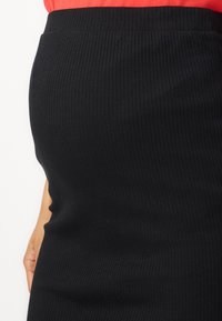 Svart, ribbet blyantkjole med en tettsittende fasong, laget av elastisk materiale. Livlinen er glatt, og teksturen har vertikale linjer.