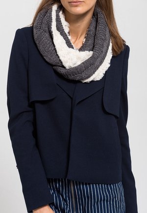 Femme portant une veste bleu marine foncé et une écharpe infinie tricotée gris-blanc, associées à un pantalon bleu à fines rayures.