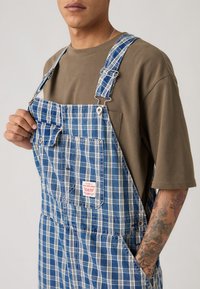 Μπλε καρό ολόσωμη φόρμα από τζιν, με ρυθμιζόμενους ιμάντες, μπροστινή τσέπη και διακοσμητική ετικέτα με λογότυπο, φορεμένη πάνω από μπεζ μπλούζα T-shirt.