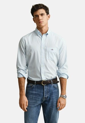 Mann trägt ein hellblau-weiß gestreiftes Hemd mit hochgekrempelten Ärmeln, blaue Jeans, geflochtenen braunen Gürtel und silberne Armbanduhr.