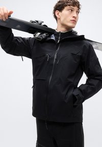 Μπουφάν για σκι σε μαύρο χρώμα, με ψηλό γιακά, πολλαπλές τσέπες με φερμουάρ και κομψό σχεδιασμό. Φαίνεται αδιάβροχο με ρυθμιζόμενους μανσέτες.