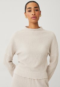 Lichtbeige pullover van gestructureerde stof. Heeft een opstaande kraag, lange mouwen en een ontspannen pasvorm, met subtiele kreukdetails overal.