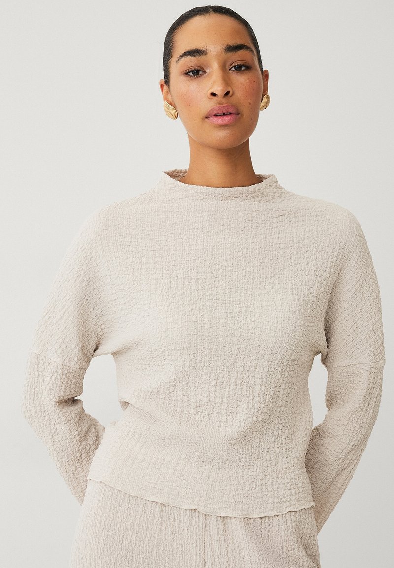 Lichtbeige pullover van gestructureerde stof. Heeft een opstaande kraag, lange mouwen en een ontspannen pasvorm, met subtiele kreukdetails overal.