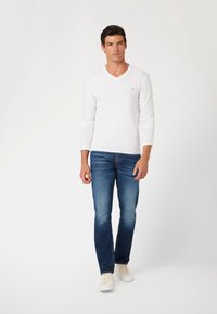 Hvid langærmet V-hals skjorte med et lille rødt logo, parret med blå denim jeans og hvide sneakers. Enkel, tætsiddende design.
